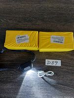 Photo showing two yellow boxed headlamp packages, one headlamp lit with white light, and a white USB charging cable, with lot number D39
