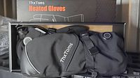 Photo showing black ThxToms heated mittens inside their packaging box with visible branding and heating control button.