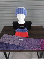 Photo showing three knit head band ear warmers in light blue, red, and dark blue, along with a long knit scarf with purple, pink and blue tones on a table, with a mannequin head displaying the light blue head band.