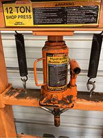 Close-up photo of 12 ton hydraulic bottle jack mounted in the center of the orange metal press frame showing label and springs