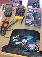 Overview photo showing two digital multimeters, an orange clamp meter, Easy Lift vehicle exit assist in original packaging, and a black zippered case with soldering iron kit