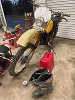 Full view of yellow dirt bike, red gas can, and black boots on concrete floor