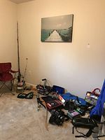 Wide shot showing folding chair, fishing rods, recurve bow, assortment of tackle items and camp stove on carpeted floor.