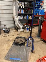 Wide view of blue floor jack, black rolling tool tray, and wrapped automotive mats in a workshop setting with automotive supplies in the background.