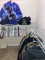 Blue zippered hooded sweatshirt with eagle graphic, folded dark work pants on wire shelf above many plastic hangers.