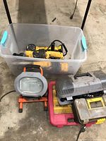 Plastic bin containing several yellow DeWalt power tools including angle grinders and sanders, with cords wrapped around and side handles visible. Surrounding the bin are multiple tool cases and a portable HELO work light on the floor.