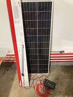 Image of solar panel leaning against wall with original packaging box visible beside it, with wires and solar charge controller unit on floor.