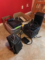 Overall lot showing Xbox 360, Sony speaker, Tzumi karaoke machine, power cords, and cables in crates and boxes on the floor.