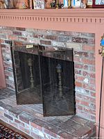 Fireplace screen set up in a brick fireplace with visible brass posts behind. The screen is black with brass hardware and four folding panels, showing the item in use.