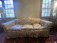 Full frontal view of antique sofa showing cream and beige floral damask upholstery, rolled arms, pleated skirt, and cushioned seating.
