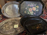 Five large vintage trays stacked and displayed, showing metal and silverplate surfaces with painted floral and bird designs.