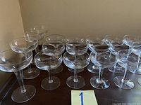 Image showing 17 small champagne coupe glasses arranged on a dark surface with light reflections on the glass.