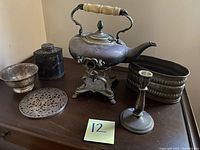 Full view of all six silverplate items arranged on a table including the teapot with ivory handle, the trivet, bowl, flask, oblong container, and candlestick.