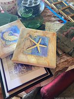 Photo showing two shell art tiles with starfish and nautilus shell designs and glassware background