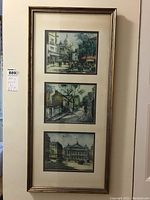 Full view of tall frame containing three European cityscape prints in vertical alignment, featuring streets with buildings, domes, and pedestrians. Frame is gold-tone with cream matting.