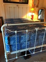 White vintage metal bedframe with blue mattress, showing decorative floral accents in the metal frame, footboard and headboard seen from a side angle.