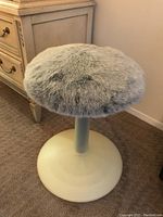 Side view of round cat perch with light gray faux fur top and cream plastic base beside wooden furniture.