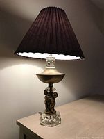 Desk lamp on table with dark pleated fabric shade, cherub figurine base on clear cut glass pedestal, bronze-colored metal parts visible.