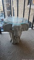 Full view of the white three-headed elephant stone side table with round glass top on balcony carpet.