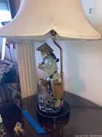 Full view of table lamp showing beige scalloped lampshade and ceramic Asian farmer figurine base with woven baskets.