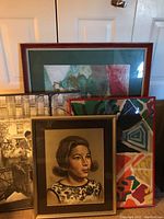 Four framed art pieces stacked against a door, including a portrait, a city street photograph, an abstract painting, and a landscape artwork.