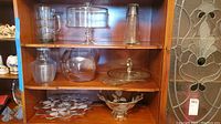 Glass serving dishes displayed on wooden shelves including pitchers, cake plate with dome, platters, bowl and vase.