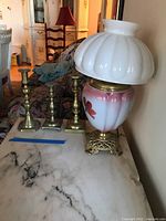 View of brass base lamp with white ribbed glass shade and three brass candlesticks on marble surface.