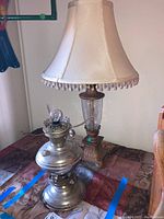 Two table lamps side by side on a table, one brass oil lamp and one electric lamp with beige fabric shade and beaded fringe