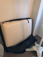 Folding massage table in beige upholstery partially inside black carrying case against a white wall.