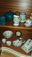 Wood cabinet shelf with collection of assorted porcelain and ceramic tea and serving items including teapot, cups, finger dishes and covered dishes.