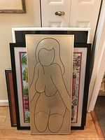 Photo showing multiple framed artworks stacked closely, foreground features metal art piece with female nude outline on silver background.