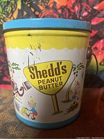 Front view of Shedd's Peanut Butter vintage metal advertising tin can showing colorful cartoon artwork and branding.