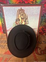 Top view of black felt hat showing rounded crown and brim with colorful poster background.