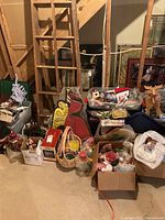 Multiple Christmas decorations and boxes including wood angel cutout, assorted baskets with ornaments, Lemax Village collectible box, and wood reindeer visible in basement setting.