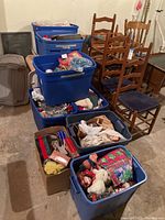 Photo showing a stack of blue bins and cardboard boxes filled with mixed Christmas decorations on floor near wooden chairs