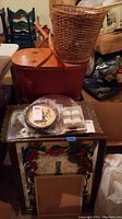 Overview photo showing the wicker picnic basket on top of a red woven basket, several framed wall art pieces including Asian-themed art, some table mirrors in plastic packaging, and other household items around them.