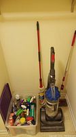 Photo showing Dyson vacuum cleaner standing next to a rope mop and small floor sweeper, along with a transparent plastic box containing assorted cleaning agents.