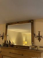 Frontal photo of large gold-framed mirror with intricate corner decorations and two brass candle sconces flanking it on the wall above mantle.