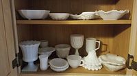 Shelf of multiple milk glass items displayed showing bowls, dishes, goblets, candle holder with hobnail pattern and scalloped edges