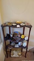 Full view of assorted items arranged on a three-tiered wooden stand including vintage ashtrays, porcelain flower, creamer pitcher, stones, glassware and small collectibles.