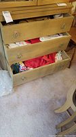 Drawers of a wooden dresser open showing various linens and small household items in soft colors including reds, whites, and patterns.
