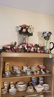 Full view of wooden shelving unit with multiple porcelain tea cups, saucers, candy dishes, sugar container on two shelves and silk floral arrangements on top