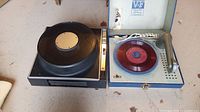 Vanity Fair turntable in an open blue textured carry case with a silver tone arm and a red RCA Victor 45 RPM record on the platter next to a black Anscorma slide projector.