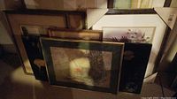 Photo showing multiple framed prints stacked against a wall, including a print with a bench and a red bird, and another print with woodland animals