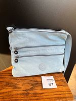 Front view of light blue Kipling Avlar cross body bag showing two zippered pockets and Kipling logo patch on wood surface.
