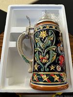 Beer stein in styrofoam packaging showing the decorated body and handle with intricate colorful relief artwork.