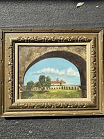 Front view of the framed Spanish style landscape painting showing the scene through an arch with building, trees, and sky.