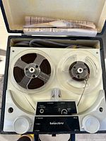 Front view of the open Telectro reel-to-reel tape recorder showing two reels with tape, control knobs, and control panel inside the case with instruction manual above.