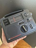 Front close-up of the black CC Solar Observer radio showing tuning dial, volume, hand crank, and speaker grille.