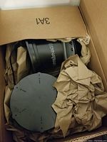 Black cylindrical mosquito trap unit inside cardboard box with kraft paper protective wrapping, showing unit and removable retaining cage.